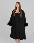 Woman wearing a long black coat with fur cuffs, standing front-facing